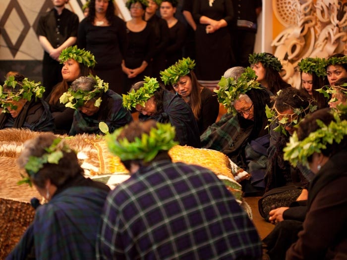 Repatriation ceremony, 2012. Photograph by Michael Hall. Te Papa Several women wearing black and wreaths on their heads made of kawakawa leaves are sitting around a cloak on some boxes in front of them.