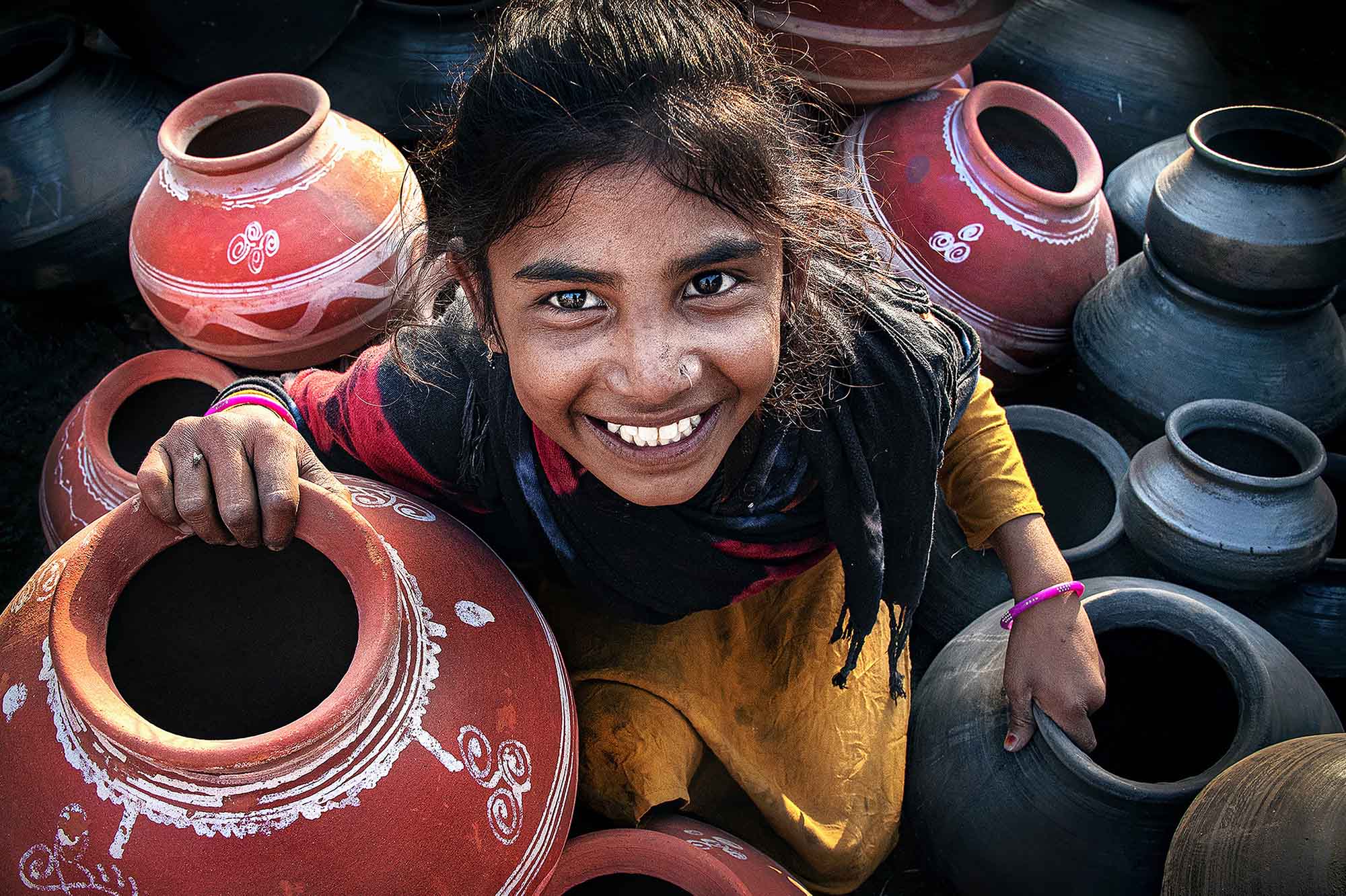 A smiling young woman sits among handcrafted earthenware pots, looking into the camera.