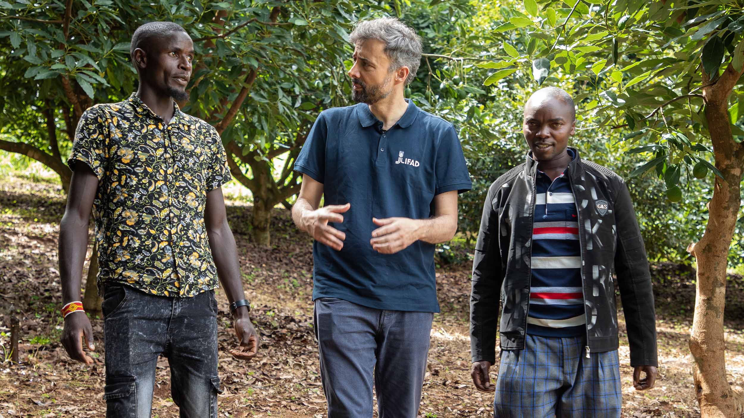Alvaro walks through dense trees with two men on each side as they chat all together