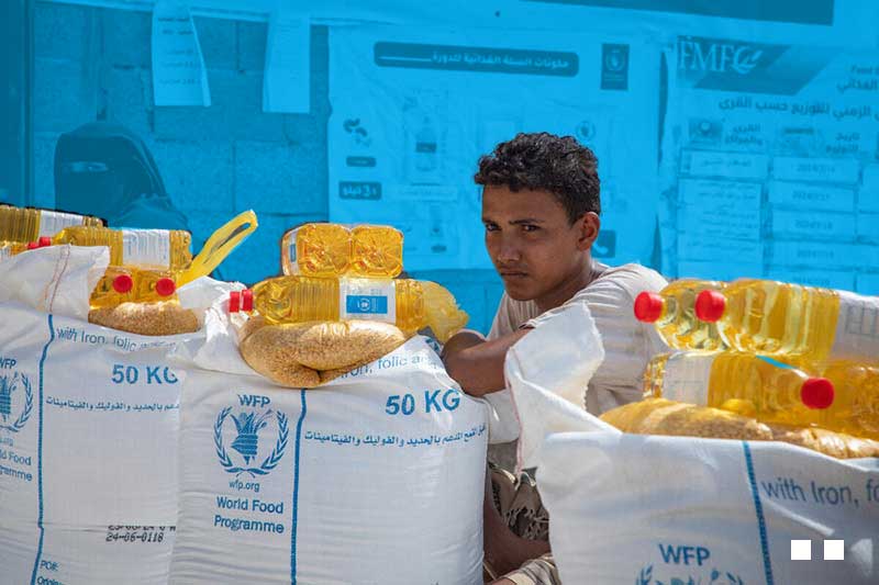 un jeune homme assis derrière des sacs de 50kg d’aide alimentaire.