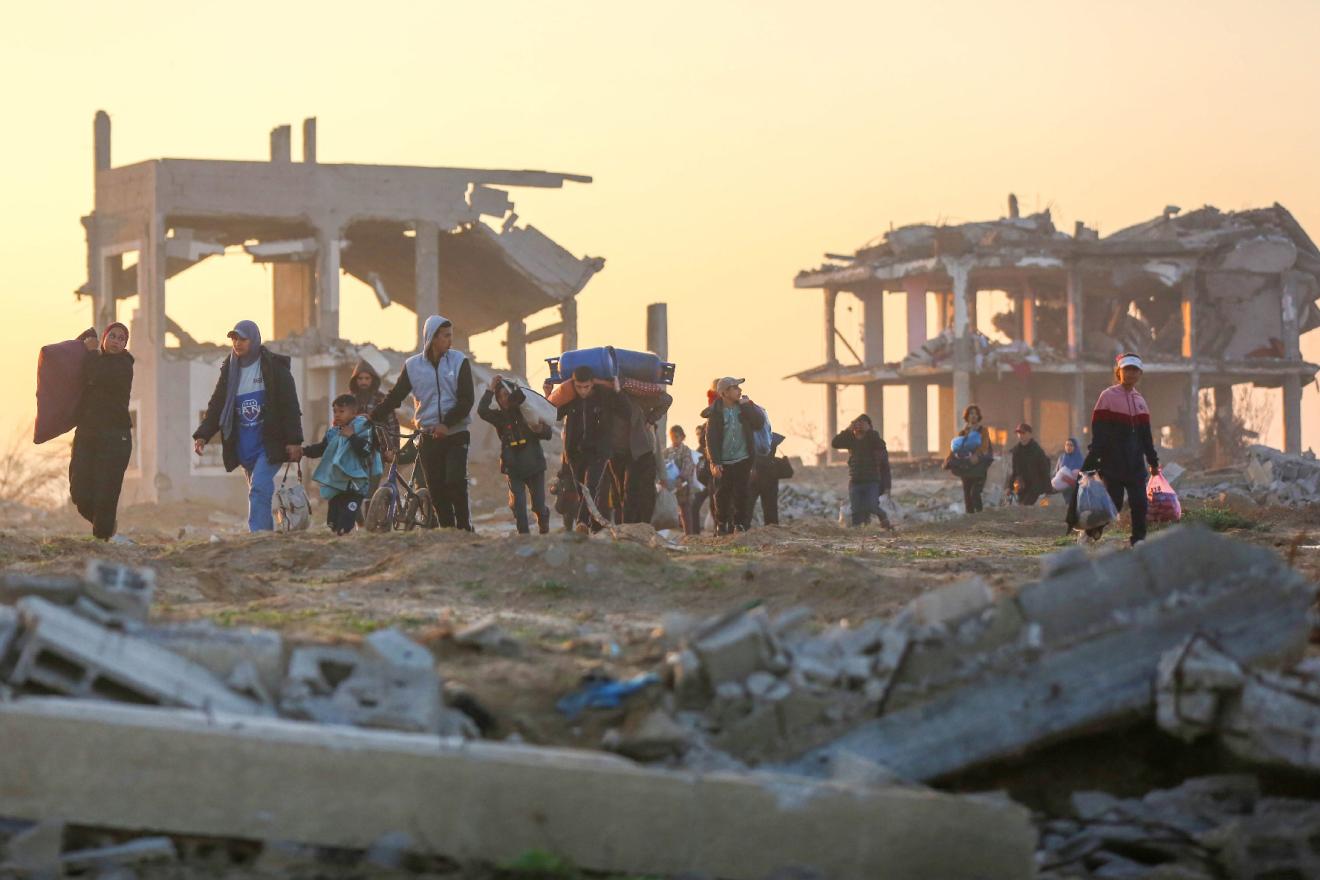 A group of people carrying their few belongings, walking in front of buildings in ruins, at sunset.