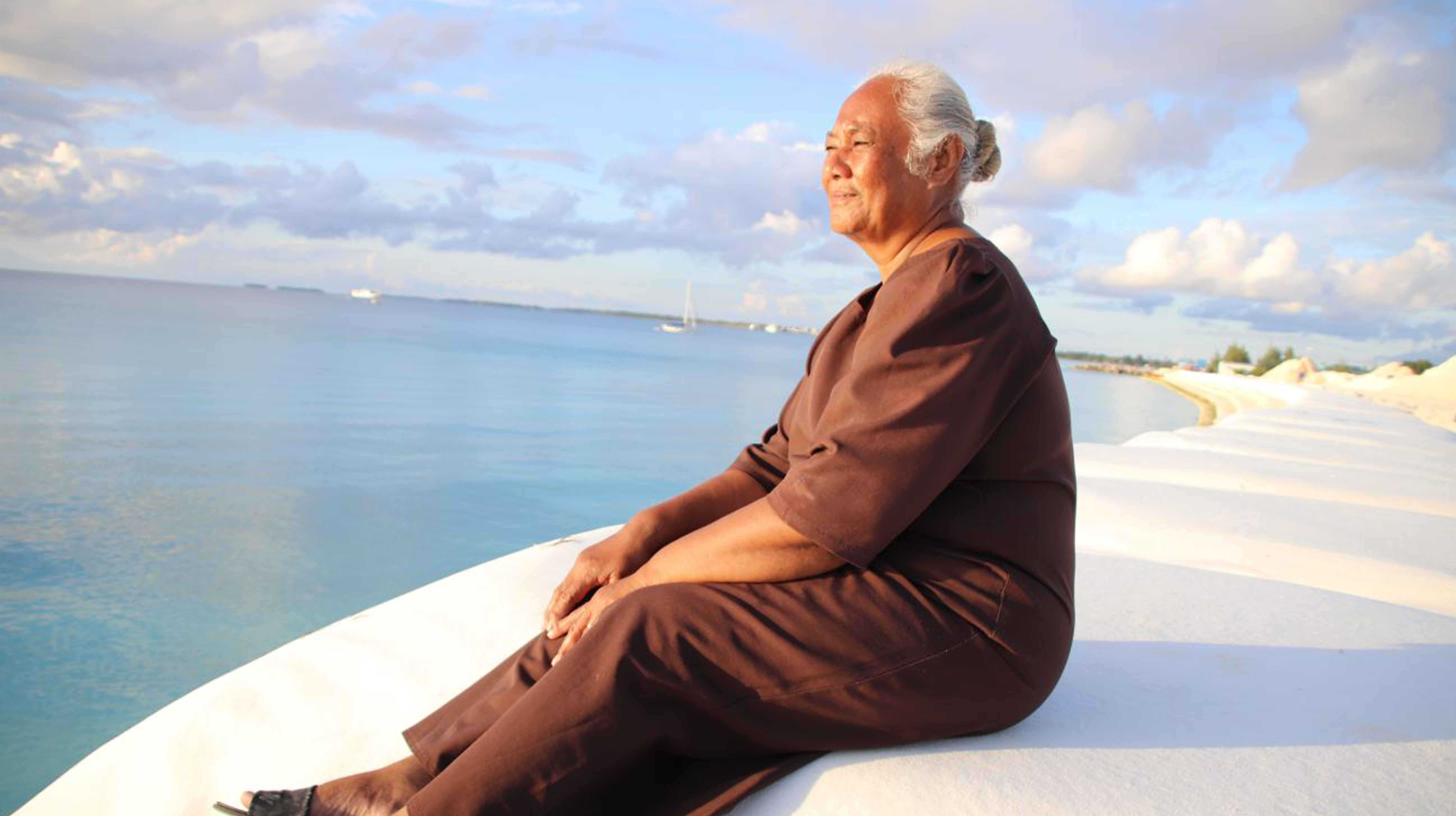 An older woman sits on a newly constructed coastal barrier overlooking the ocean in Tuvalu