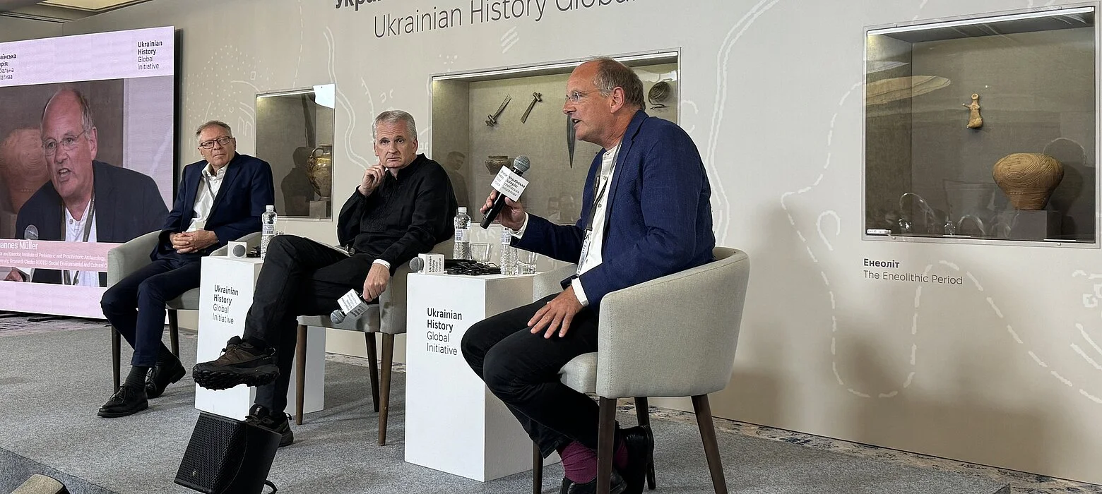Three men are sitting on a podium. Behind them, there are display cases with archaeological finds on the wall.
