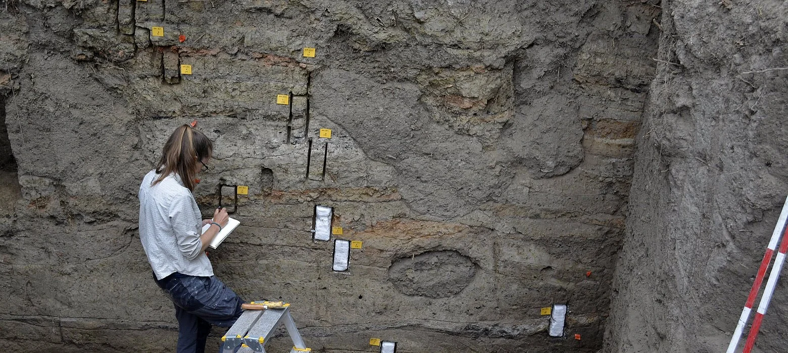 A woman is standing in an excavation trench that is at least three meters deep. Various layers are marked with labels in the profile in front of her.