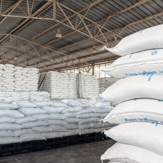 A warehouse with rows of white sacks of food stacked high on black pallets under a metal roof supported by a framework of beams.