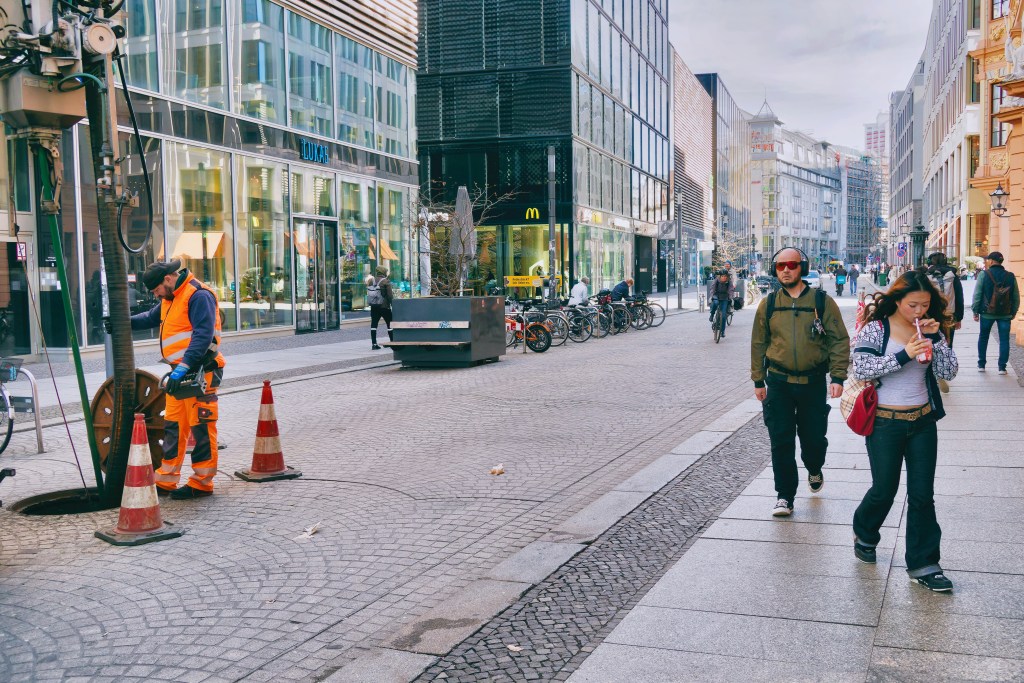 An everyday street scene in Leipzig. A girl is drinking from a cup using a straw. Next to her, a city worker is standing by a vehicle, cleaning a storm drain with a hose. The photo is a street photograph.