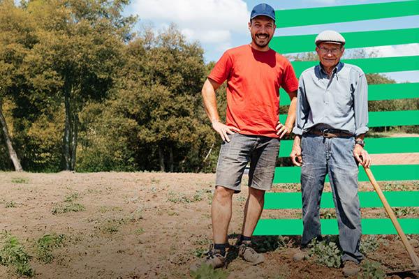 Two men stand in a field bordered by trees, the older man is holding a hoe.