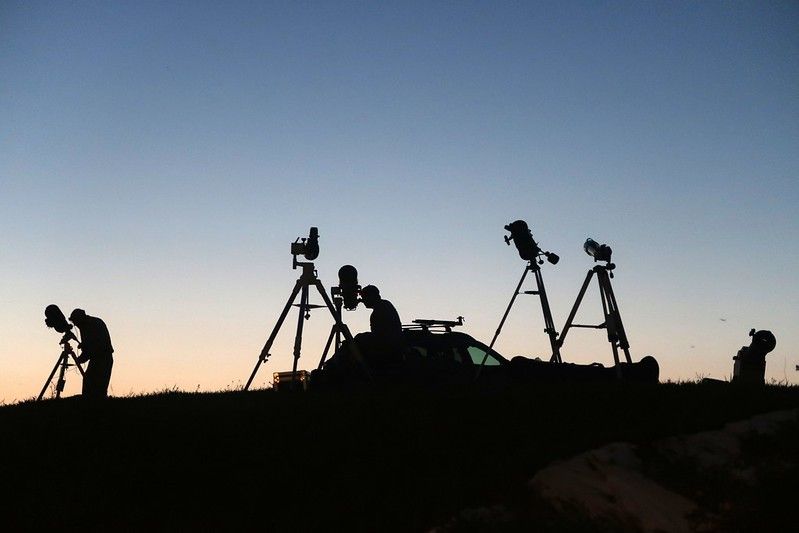 Siluetas de un grupo de personas que observan el cielo con telescopios montados en trípodes durante el crepúsculo.