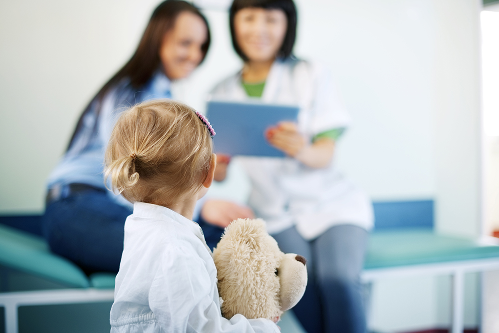 A child on his back holding a teddy bear. In the background is a woman sitting next to a health professional. They are sitting and looking at a clipboard that the health worker is holding. A child on his back holding a teddy bear. In the background is a woman sitting next to a health professional. They are sitting and looking at a clipboard that the health worker is holding.