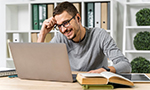 The Open University of Brazil in the management of student permanence in Pedagogy courses Photograph of a man wearing glasses and headphones smiling while looking at a laptop screen. He is sitting at a table with a book, notebooks and a pen. In the background, there are shelves with folders and potted plants.