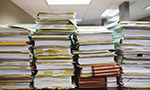 Fotografía que muestra cuatro pilas de documentos que ocupan casi todo el espacio de la imagen. Al fondo se ve el techo.