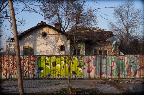 A Long-Vanished Nightspot: A patch of pavement, a corrugated metal fence, and a rundown cottage on a main thoroughfare in Sofia, Bulgaria – the likely location of a nightclub once owned and run by Keva, a legendary Romani (Gypsy) vocalist in the years preceding the Second World War. In its day, Cafe Keva was a popular gathering-place for Sofia residents of diverse ethnicities and walks of life. Fuji X100, 2014. Click on image to enlarge.