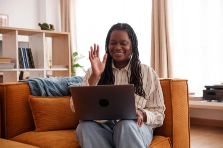 A woman with long, braided hair smiles and waves while sitting on a couch, using a laptop in a cozy living room