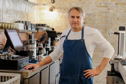 A middle-aged man in a denim apron stands confidently at a café counter, with coffee equipment and wine glasses visible in the background
