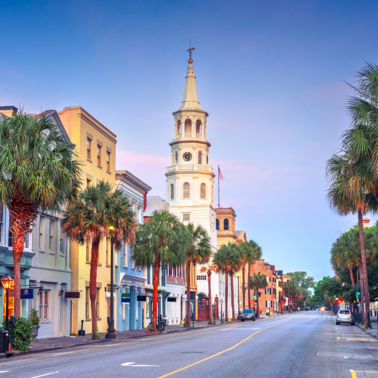 A wide street lined with palm trees, featuring historic buildings in Charleston, South Carolina. The steeple of a church is prominent against a pastel sky during dawn.