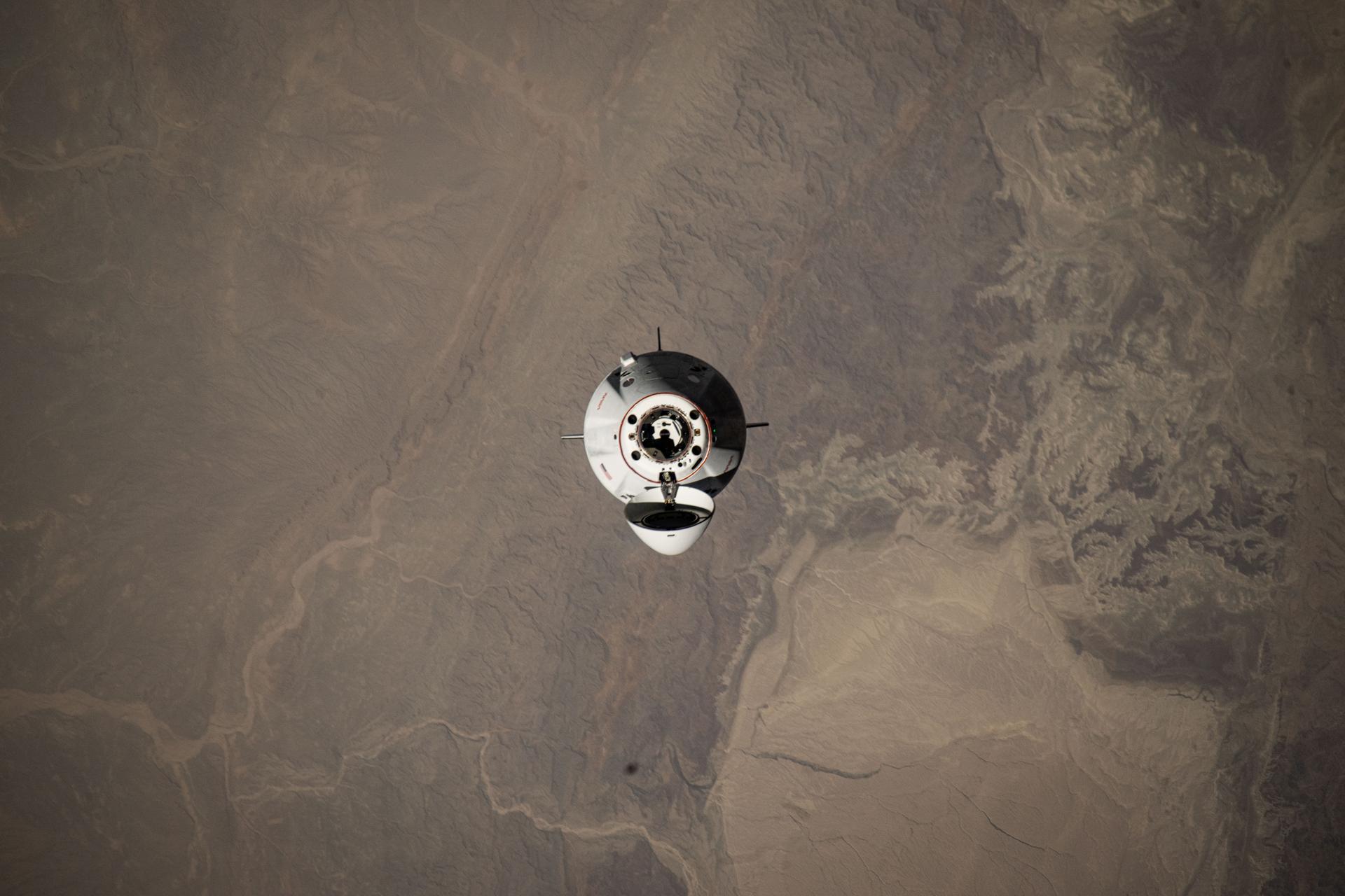 A white space capsule is seen floating above the brown surface of Earth