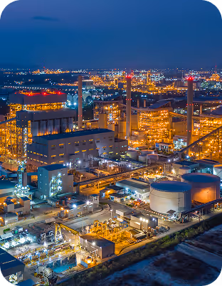 Industrieanlage mit mehreren beleuchteten Gebäuden und Lagertanks bei Nacht unter einem dunkelblauen Himmel.