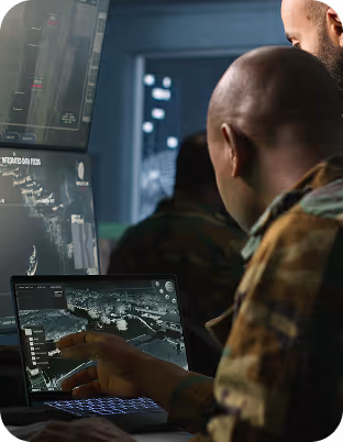 Zwei Männer in Tarnuniformen arbeiten an einem Laptop, auf dem eine detaillierte Kartenoberfläche in einem Kontrollraum angezeigt wird.