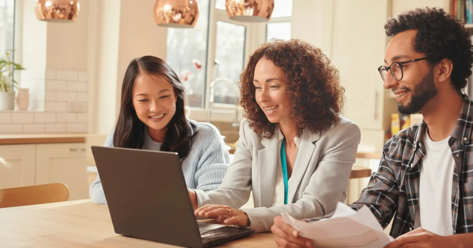 Three people sitting at a table in a kitchen, one using a laptop while another holds papers, all smiling.