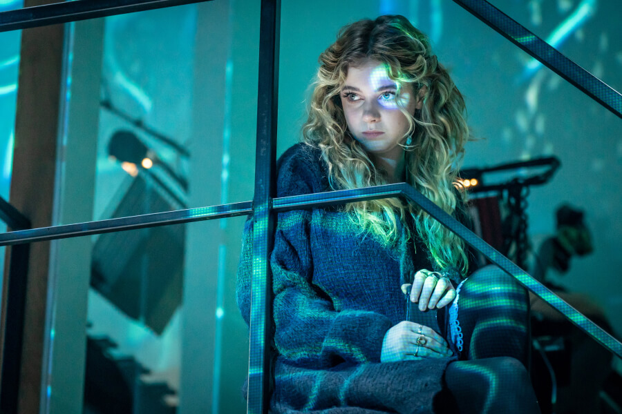 Natalie, played by Eleanor Worthington-Cox, sits pensively on a staircase, illuminated by blue and green lights. She has long, wavy blonde hair and is wearing a dark, oversized sweater. She has a thoughtful, distant expression on her face. The setting appears to be modern and slightly industrial, with metal railings and a blurred background that includes various objects, creating a moody and introspective atmosphere.