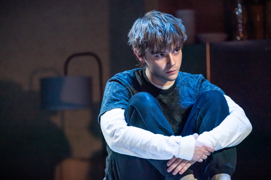 Gabe, played by Jack Wolfe, sits on the floor, hugging his knees to his chest. He is dressed in a layered outfit with a long-sleeve shirt under a t-shirt, and he has a serious, contemplative expression on his face. The background is dimly lit, with a blurred lamp and other indistinct objects, creating a somber and introspective mood.