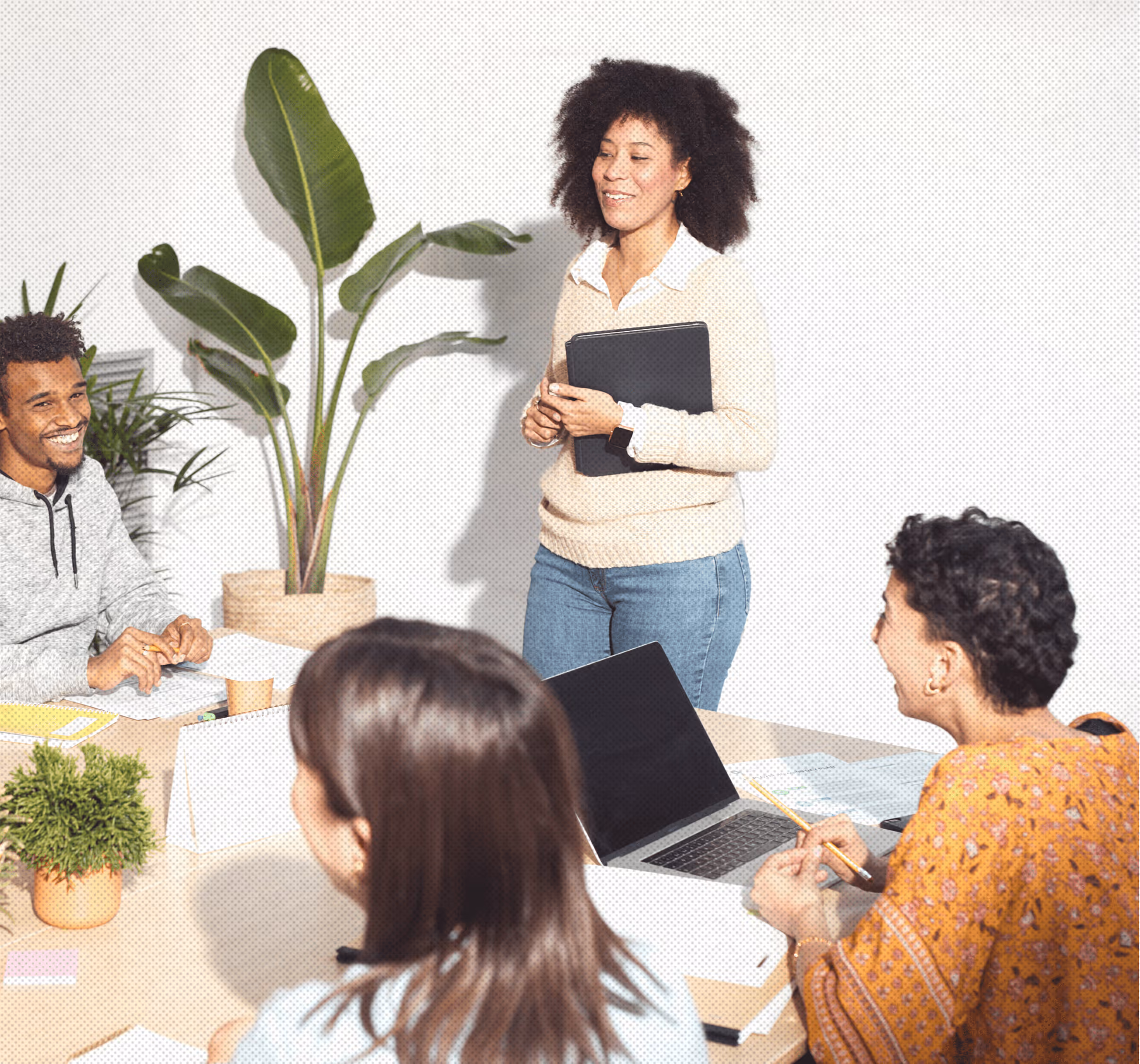Four diverse coworkers engaged in a meeting with one standing woman holding a folder, smiling colleagues seated around a table with a laptop and plants.