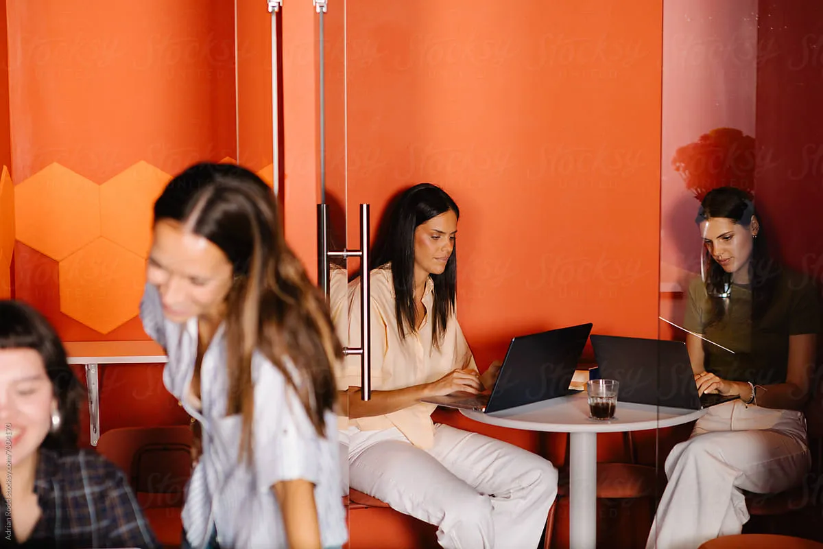 Woman working on a laptop at a table with a glass of drink in an orange-walled office space.