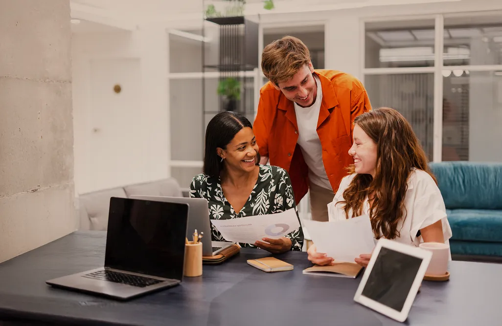Trois collègues souriants et examinant des documents ensemble à un bureau avec des ordinateurs portables et une tablette.