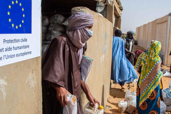 A woman leaving a house with goods in her hands. Outside on the street children and other women holding goods as well.
