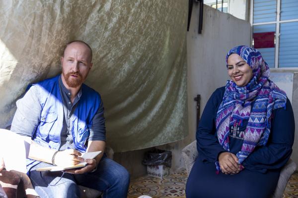 A woman wearing a colourful patterned headscarf and dark clothing sits on a chair, with hands clasped, while another man in a blue shirt writes on a notepad in a sunlit room. The background includes a patterned floor, a window with blue panels, and a light-coloured curtain.