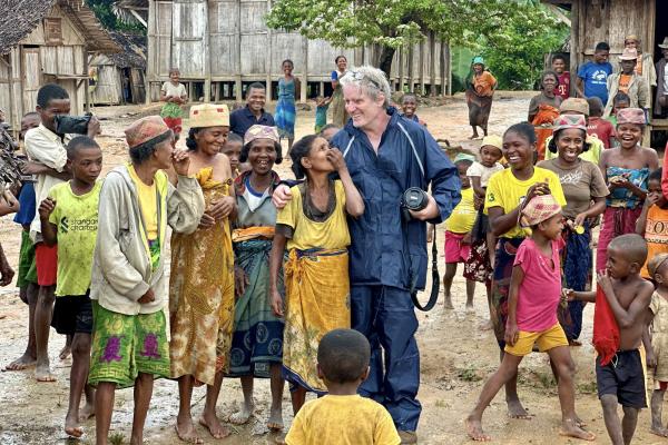 A group of locals, including women and children dressed in colourful traditional clothing, gathered outdoors in a rural setting with wooden houses in the background. A photographer is holding a camera and standing among them, engaging with the group.
