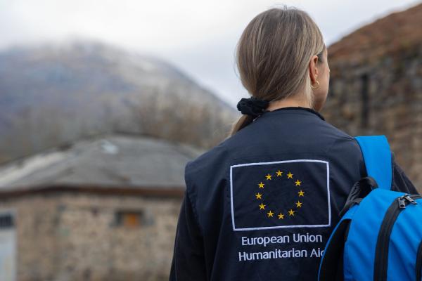 A humanitarian aid worker wearing a jacket with the European Union Humanitarian Aid logo on the back, standing outdoors with a blue backpack. The background shows a rural setting with stone buildings and mountains under a cloudy sky.