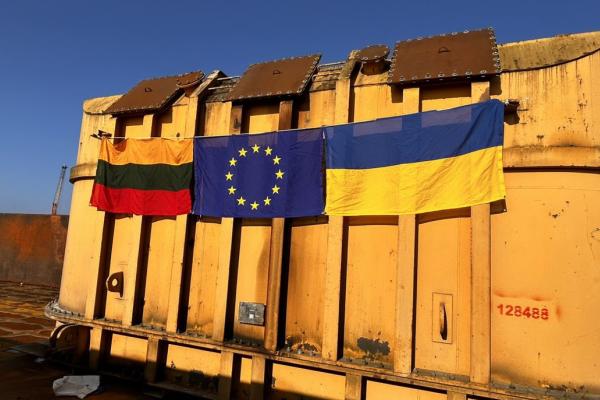 3 flags are displayed on a large yellow industrial transformer: the Lithuanian flag, the European Union flag, and the Ukrainian flag. The setting appears to be a port area under a clear blue sky.