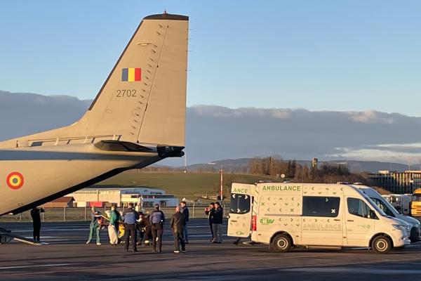 A military aircraft with the Romanian flag on its tail is parked on an airstrip, with medical personnel and law enforcement officers assisting in the transfer of individuals to a nearby ambulance marked with "CHUV". The scene takes place under a clear sky with a backdrop of distant hills and buildings.