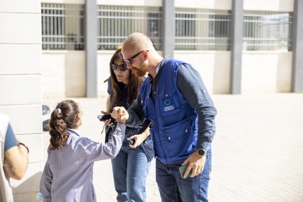 A young girl greets a man and a woman in blue vests with the European Union emblem. The setting appears to be an outdoor area of the school with barred windows.