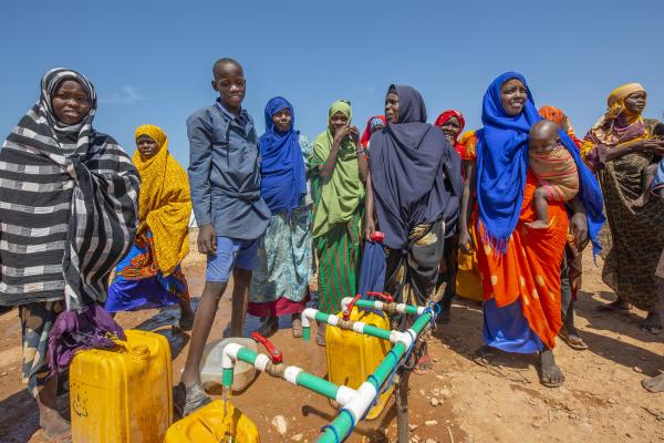 A group of people, including women in brightly coloured headscarves and long dresses and several children, stand outdoors on dry, dusty ground under a clear blue sky in Somalia. They are gathered around a simple tap stand made from plastic pipes, filling large yellow jerrycans with clean water.
