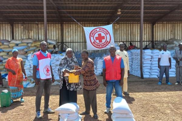 Volunteers from the Burkina Faso Red Cross distribute humanitarian aid in front of a large open-sided warehouse stacked with white sacks of food or supplies. Several people stand in a line while one person hands a yellow container or package to another in the centre. A Red Cross Burkina Faso banner hangs above them, and additional piles of filled sacks are arranged on the ground in the foreground.