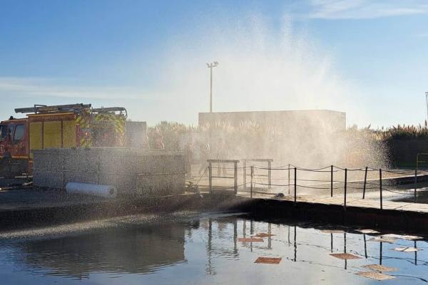 A fire engine is parked beside an industrial-style waterfront area while a powerful spray of water arches into the air, creating a misty cloud in bright sunlight. In the foreground, calm water reflects the sky and structures, with several floating pads or booms and a small white absorbent bundle suggesting a marine pollution or spill-response exercise. Low concrete walls, safety railings, and distant vegetation and buildings frame the scene under a clear blue sky.