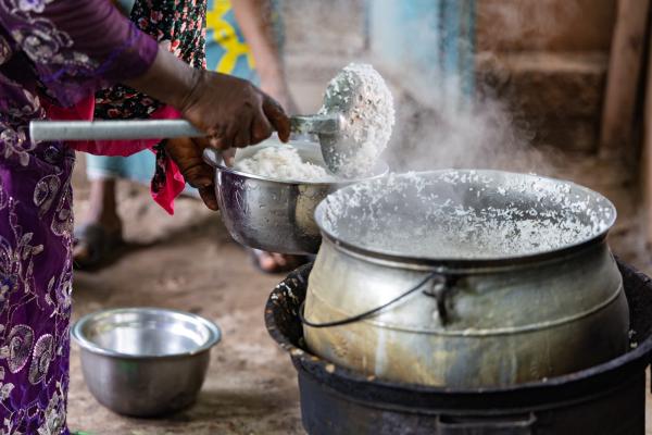 A person in brightly patterned clothing ladles steaming cooked rice from a large metal pot over an open stove into a smaller bowl, suggesting a simple communal meal in a setting with limited resources.