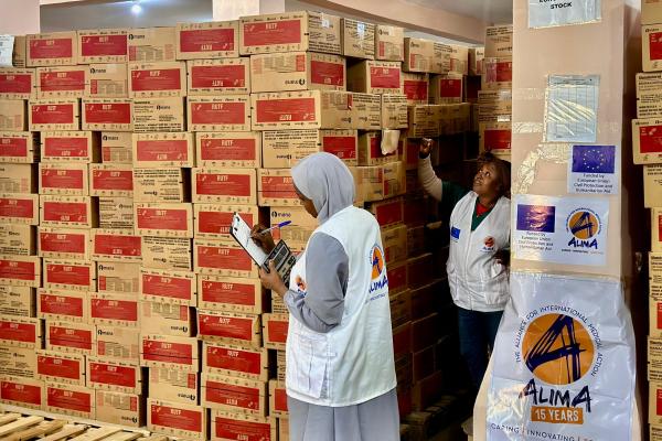Inside a warehouse, two aid workers wearing white vests with the ALIMA (The Alliance for International Medical Action) logo check inventory against a clipboard, surrounded by tall stacks of cardboard cartons labelled “RUTF” (ready‑to‑use therapeutic food). Signs on a pillar show the European Union flag and text indicating that the stock is funded by EU civil protection and humanitarian aid.