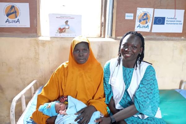 A woman in orange clothes sits on a bed with a newborn in a blue blanket, with a smiling healthcare worker beside them.