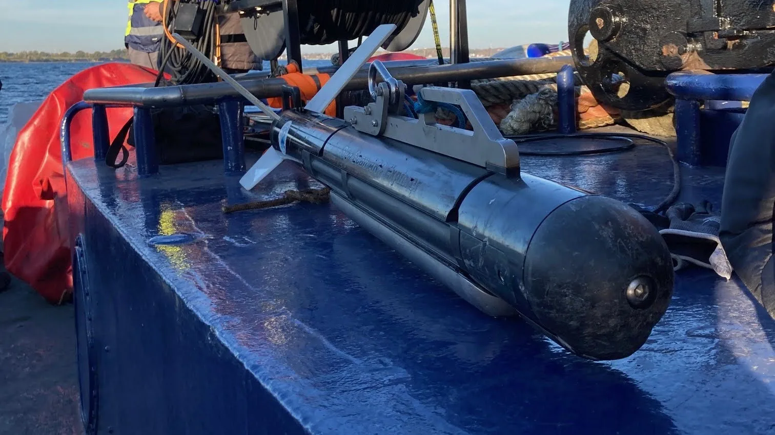 A black sonar device that looks like a torpedo sits on a metal box of a boat, with water in the background showing the boat is out at sea.