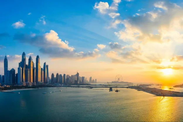 Image of a city skyline on the water with sky, clouds, and sunset.
