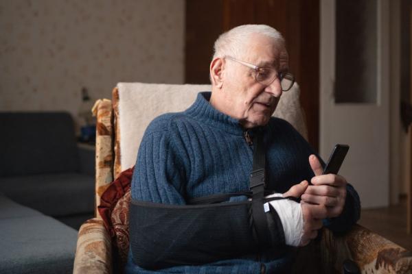 Senior with injured arm in sling browsing smartphone.