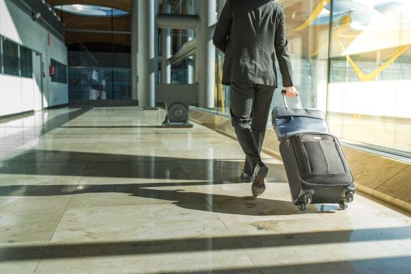 A person in a suit walks away from the camera, pulling a wheeled suitcase through a modern building with large windows.