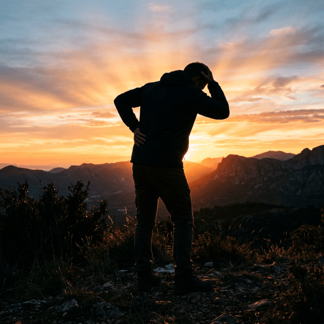 Silhouette of a person standing on rocky mountain terrain with hands on head and back at sunset