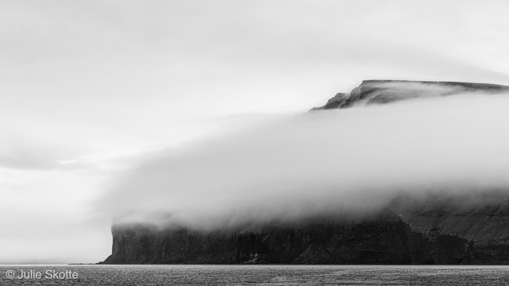 Boat trip around Svinoy og Fugloy in the Faroe Islands with low hanging clouds around the rugged landcapes. The image is in black and white.
