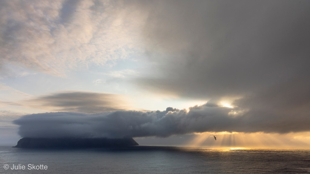 Mykines island (in the Faroe Islands) covered in clouds and sun rays through the clouds over the ocean.