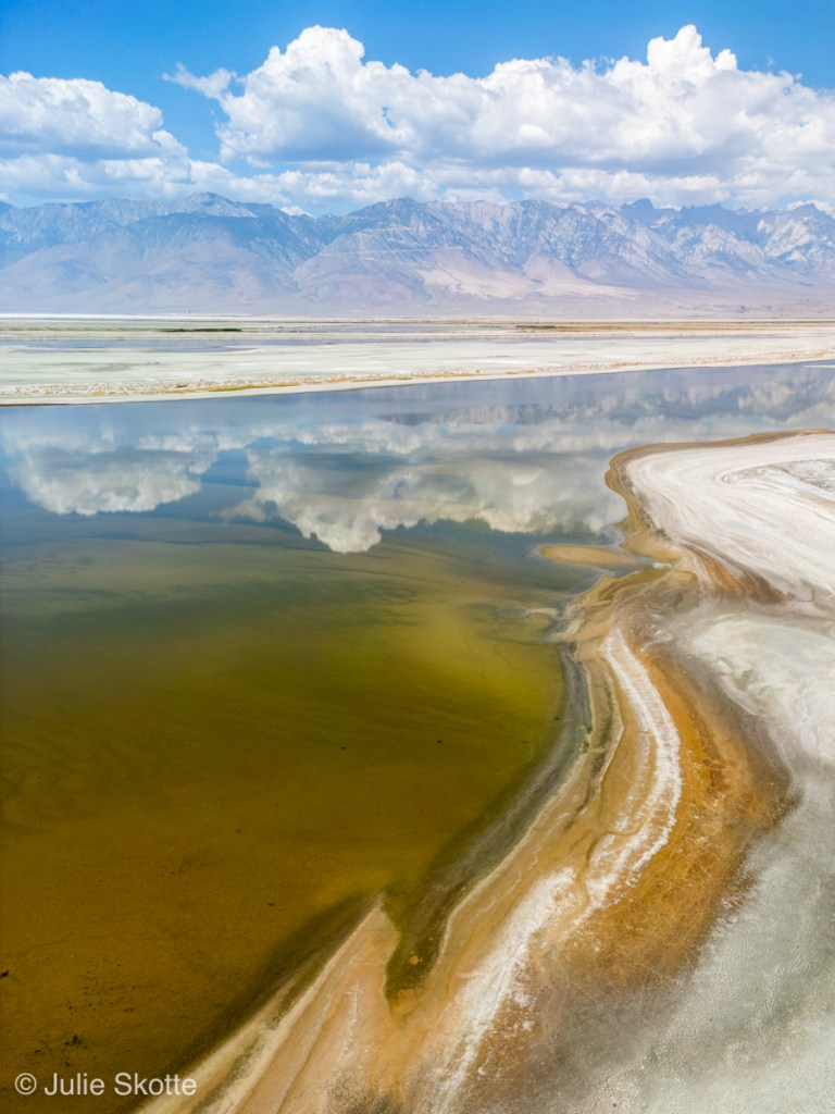 Drone billede af Owens Lake i Californien, USA. I baggrunden ses Sierra Nevada bjergene med skyer over bjergene, og dette spejles i det stille vand i Owens Lake.