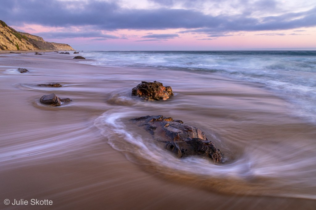 Sunset at Crystal Cove State Park in California. The sky is colored purple and pink. The waves retreats on the sand and rocks, forming lines.